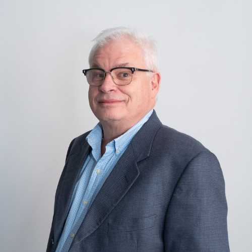 Older man with gray hair and glasses in a blue jacket, smiling—looks like a trusted CPA standing against a plain light background. - kotsoconsulting.com Todd Breckel, CPA